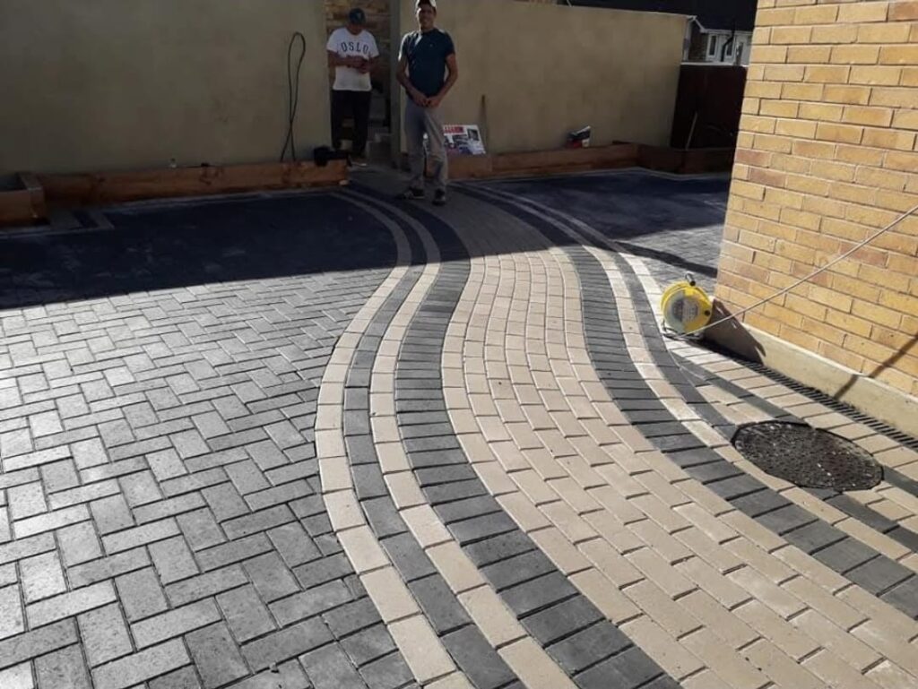 Interlocking block paving driveway installation featuring a striking curved design with contrasting grey and cream blocks, herringbone patterns, and two contractors standing in the background
