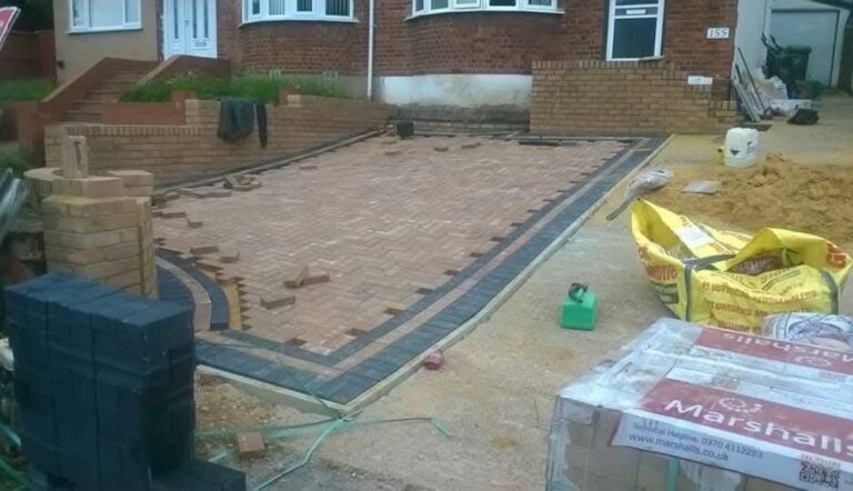 In-progress installation of a herringbone-pattern block paving driveway featuring contrasting dark borders, brick retaining walls, and stacked paving materials in front of a residential home