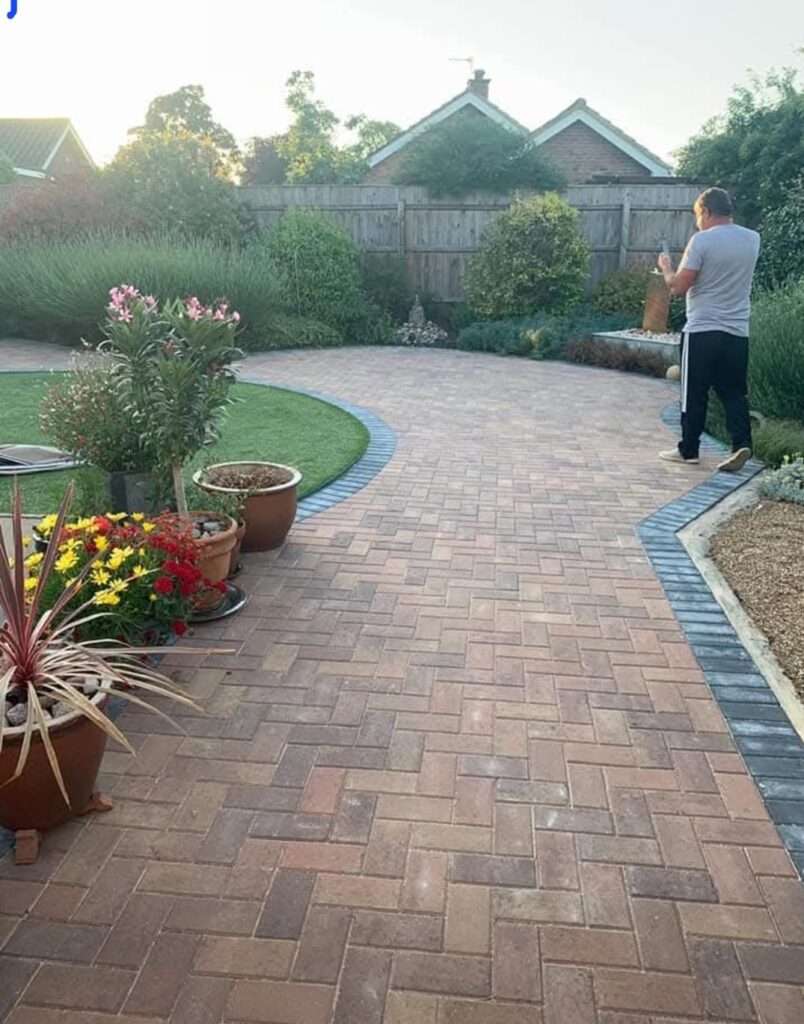 Extensive brindle concrete block paving driveway laid in a herringbone pattern with a dark contrasting soldier course border, curved lawn edge, and potted plants