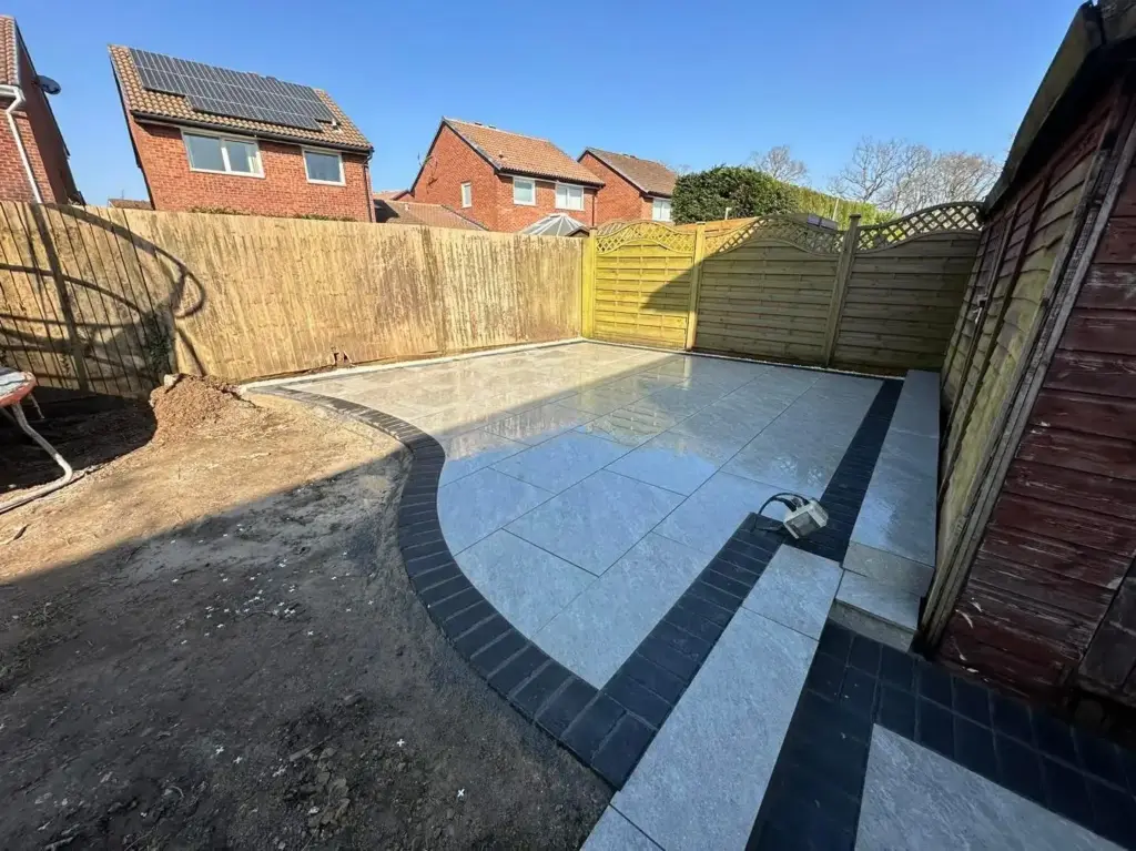 Grey porcelain slab patio featuring a sweeping curved dark soldier course border, a low raised step, and a decorative gravel strip against a wooden shed and panel fencing
