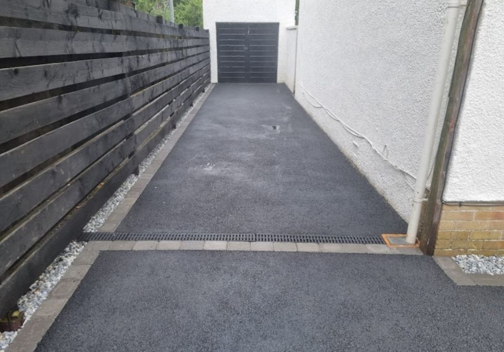 Smooth black tarmac driveway installation with grey block paving edging, across-drive channel drainage grate, dark horizontal timber fence, and white rendered house side wall leading to a garage