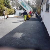 Landscaping crew operating a roller compactor on a newly laid smooth asphalt driveway with an integrated drainage cover and a delivery lorry in the background
