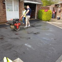 Landscaping contractors using a ride-on roller to compact a freshly laid black asphalt driveway outside a residential brick home with a garage