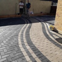Interlocking block paving driveway installation featuring a striking curved design with contrasting grey and cream blocks, herringbone patterns, and two contractors standing in the background