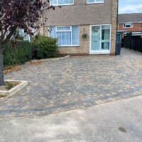 Multi-blend block paving driveway installed in a herringbone pattern with charcoal block border edging, raised stone garden beds, and front porch entrance