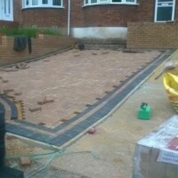 In-progress installation of a herringbone-pattern block paving driveway featuring contrasting dark borders, brick retaining walls, and stacked paving materials in front of a residential home