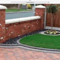 Red brick boundary wall with matching pillars and stone copings, overlooking a front lawn with black slate chippings, a circular flower bed, and a block-paved driveway