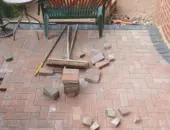 Red brick weave block paving patio under construction featuring a dark soldier course border, stacked pavers, a stiff broom, and a brick retaining wall