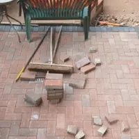 Red brick weave block paving patio under construction featuring a dark soldier course border, stacked pavers, a stiff broom, and a brick retaining wall