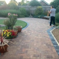 Extensive brindle concrete block paving driveway laid in a herringbone pattern with a dark contrasting soldier course border, curved lawn edge, and potted plants
