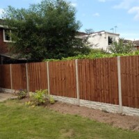 Newly stained brown wooden lap fence panels fitted between durable concrete posts and textured patterned concrete gravel boards, bordering a residential lawn and garden bed
