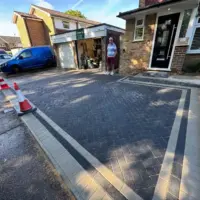 Completed dark charcoal block paving driveway in a herringbone layout with double contrasting stretcher bond borders, leading to the entrance of a semi-detached brick home with a modern black front door