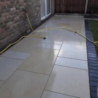 Wet cream sandstone patio stretching along a traditional brick home exterior, featuring a dark brick soldier course border, a green lawn, and a coiled yellow garden hose