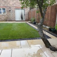 Cream sandstone patio featuring precision wet-cut joints and a mature tree cutout, bordered by a dark brick soldier course edging, a freshly laid turf lawn, and wooden fencing