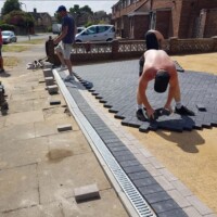 Landscaping contractors installing a dark grey block paving driveway in a herringbone pattern with integrated linear drainage channels and a sub-base preparation layer