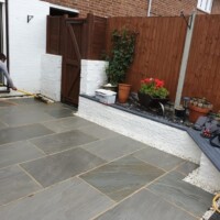 Landscaping contractor sweeping joint compound into a newly laid dark grey sandstone flagstone patio, bordered by white-rendered raised walls, potted plants, and a wooden fence