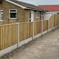 Stepped featheredge wooden fencing with concrete posts and gravel boards running alongside a concrete driveway and a residential yellow-brick bungalow