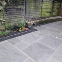 Grey sandstone flagstone patio featuring a dark brick soldier course border, integrated drainage, and an adjacent garden flower bed enclosed by wooden fence panels