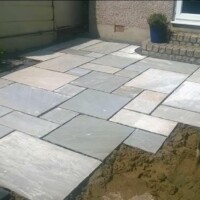 Newly installed light grey sandstone flagstone patio with dark mortar joints, adjoining a brick patio step and a garden excavation area with a shovel