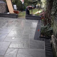 Professional grey sandstone patio installation featuring crisp dark brick soldier course borders, integrated drainage, raised brick planters, and a landscaper working in the background