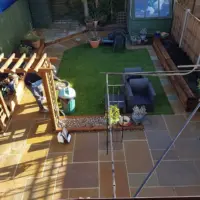 Professionally landscaped back garden featuring rich brown sandstone flagstone paving, a central green turf lawn, timber sleeper raised beds, a wooden pergola frame, and a greenhouse