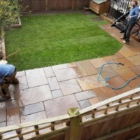 Landscaping contractors installing a rich brown sandstone flagstone patio and a newly laid green turf lawn, framed by timber sleeper borders and wooden fence panels