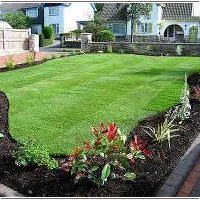 Immaculately manicured front lawn featuring curved bark-mulched flower beds with vibrant plants, bordered by a neat block-paving driveway and a residential suburban home