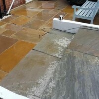 Multi-level patio installation featuring warm yellow-brown and dark grey sandstone flagstones, recessed manhole covers, white-rendered retaining walls, and an adjoining garden lawn