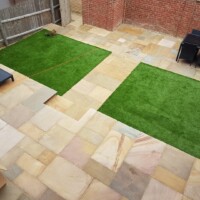 Contemporary multi-level garden design featuring varied sandstone flagstone paving, integrated artificial grass lawn sections, outdoor patio furniture, and traditional brick retaining walls