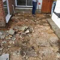 Landscaping contractors breaking up old concrete paving and excavating a patio sub-base alongside a brick wall and patio doors prior to new installation