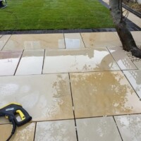 Pressure washing a wet, light-yellow sandstone flagstone patio using a high-pressure lance, bordered by a dark set-block edging and a manicured green lawn with a small tree