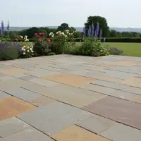 Multi-tone Rajah blend Indian sandstone patio laid in a random coursed pattern, bordered by vibrant lavender, delphiniums, and roses, overlooking a sprawling countryside lawn and distant hills