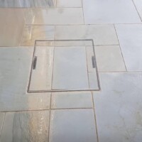 Precision-fitted recessed manhole cover seamlessly integrated into a wet light-grey sandstone patio with neat pointing and flush flagstone alignment