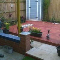 Multi-level garden featuring a freshly stained red wooden decking area, flagstone paving steps, low brick planters, potted plants, and wooden lap fencing with a grey storage shed