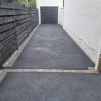 Smooth black tarmac driveway installation with grey block paving edging, across-drive channel drainage grate, dark horizontal timber fence, and white rendered house side wall leading to a garage