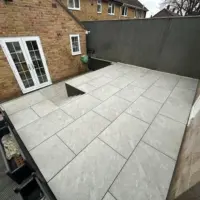 Textured light grey porcelain paving patio featuring integrated brick steps and drainage channels, set against a multi-toned brick house with white French doors and dark vertical fencing