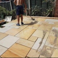 Landscaping contractor brushing jointing compound into a newly laid yellow sandstone flagstone patio, bordered by a brick edge and wooden fencing