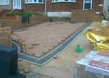 In-progress installation of a herringbone-pattern block paving driveway featuring contrasting dark borders, brick retaining walls, and stacked paving materials in front of a residential home