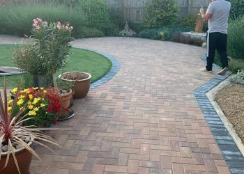 Extensive brindle concrete block paving driveway laid in a herringbone pattern with a dark contrasting soldier course border, curved lawn edge, and potted plants