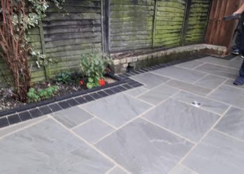 Grey sandstone flagstone patio featuring a dark brick soldier course border, integrated drainage, and an adjacent garden flower bed enclosed by wooden fence panels