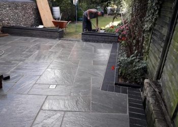 Professional grey sandstone patio installation featuring crisp dark brick soldier course borders, integrated drainage, raised brick planters, and a landscaper working in the background