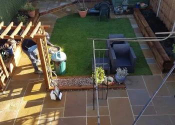Professionally landscaped back garden featuring rich brown sandstone flagstone paving, a central green turf lawn, timber sleeper raised beds, a wooden pergola frame, and a greenhouse