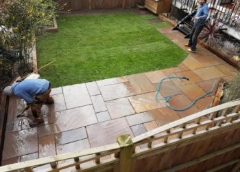 Landscaping contractors installing a rich brown sandstone flagstone patio and a newly laid green turf lawn, framed by timber sleeper borders and wooden fence panels