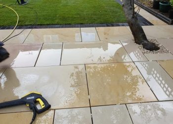 Pressure washing a wet, light-yellow sandstone flagstone patio using a high-pressure lance, bordered by a dark set-block edging and a manicured green lawn with a small tree