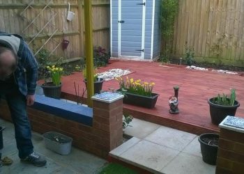 Multi-level garden featuring a freshly stained red wooden decking area, flagstone paving steps, low brick planters, potted plants, and wooden lap fencing with a grey storage shed